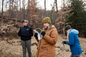 A group reviews the Lowries Run Slopes Expansion Project Land. Three explorers are in coats in the foreground on an early spring day looking at maps while hiking through trees.